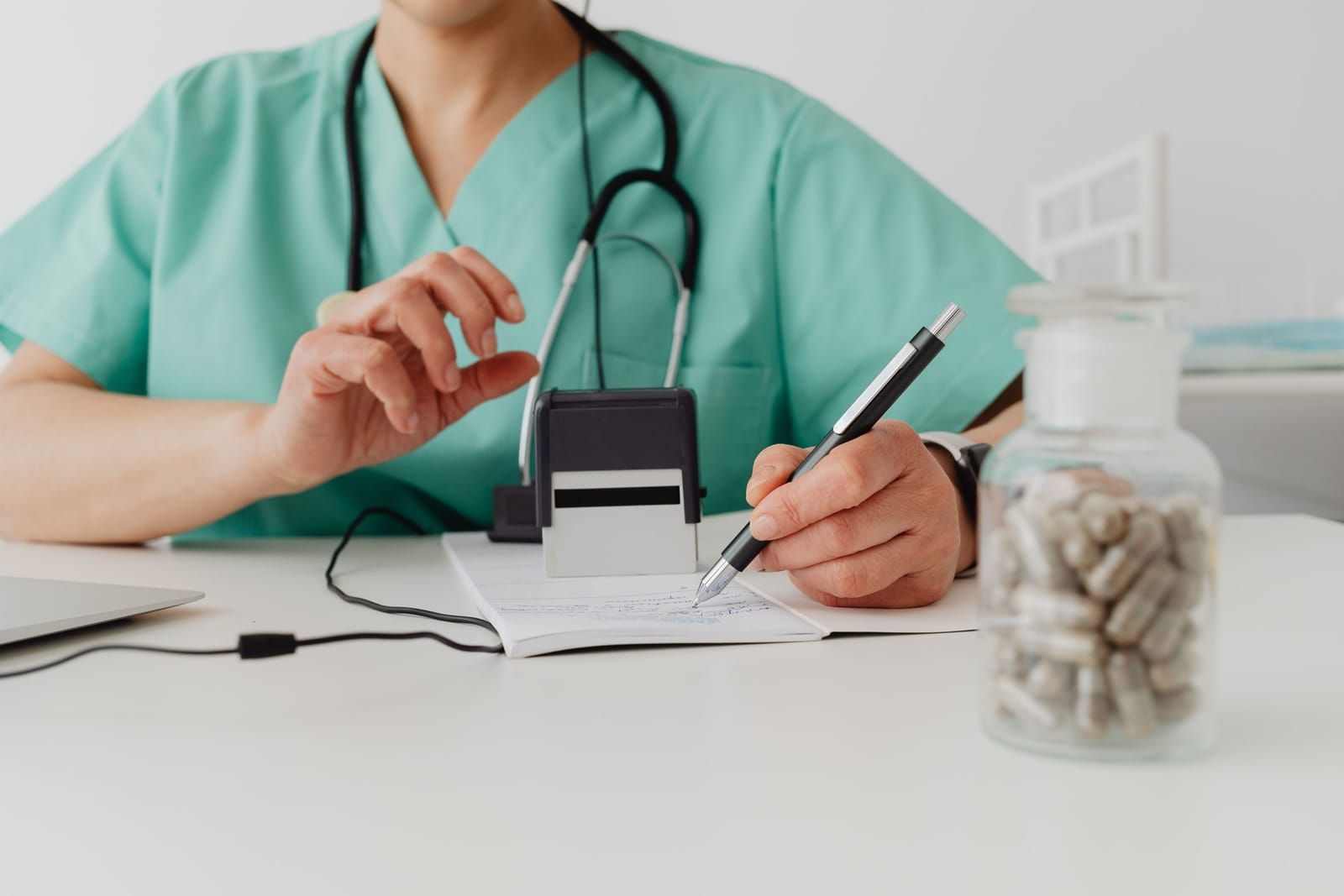 A clinician in teal scrubs with a stethoscope writing on a prescription pad, a self-inking prescriber stamp and a glass jar of capsules on the white desk beside her
