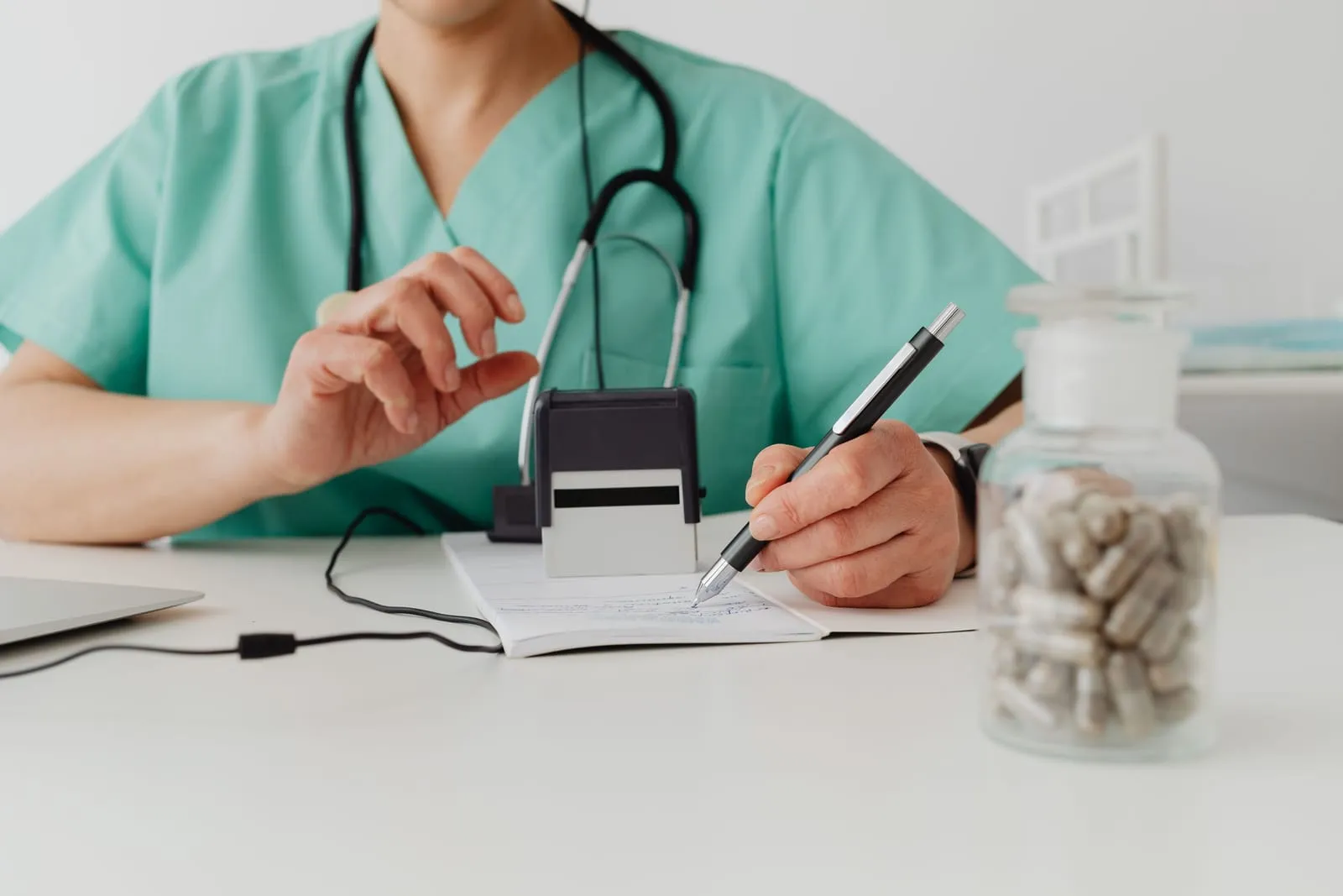 A clinician in green scrubs writing notes at a desk beside a stethoscope