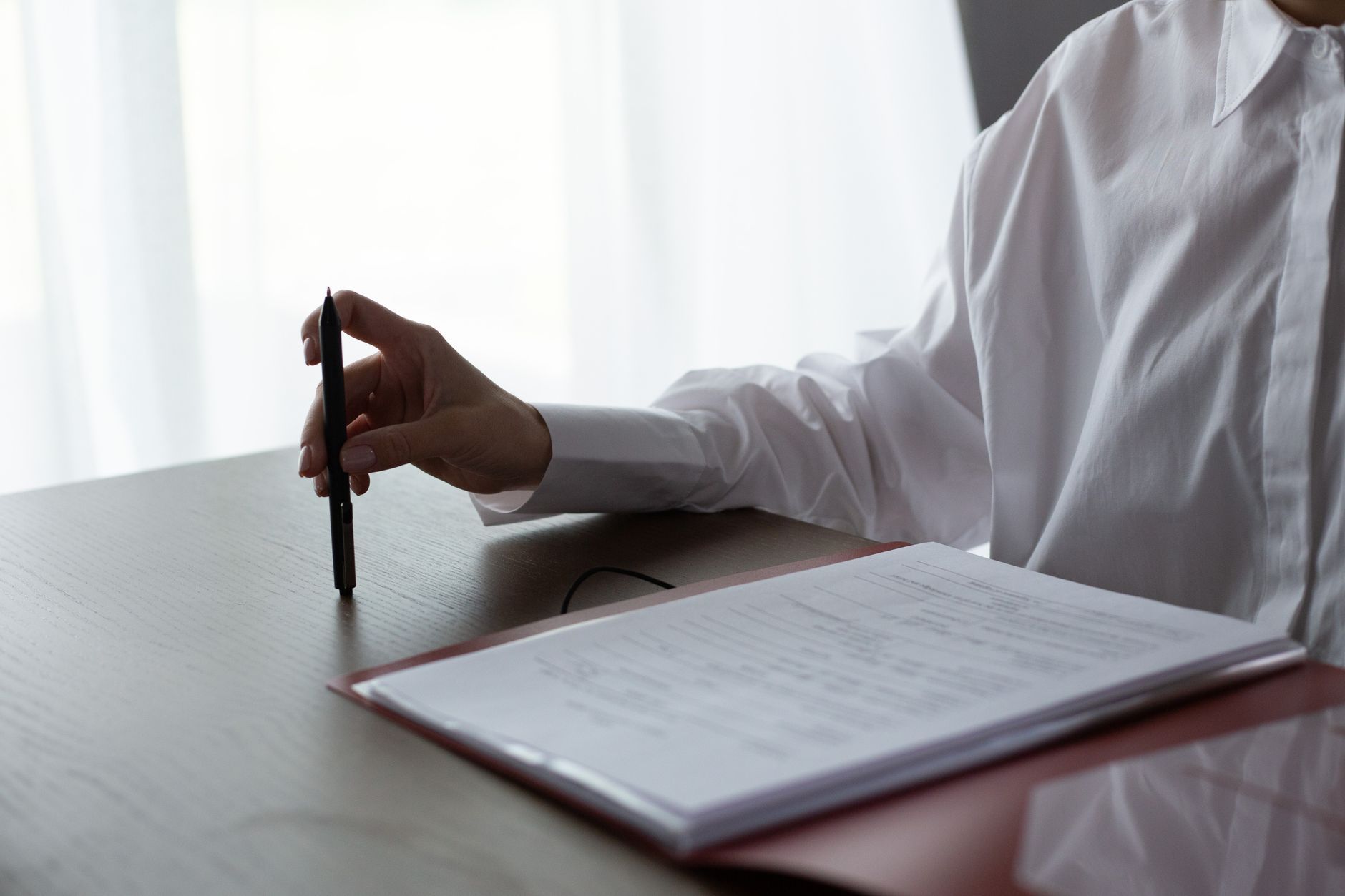A person in a white shirt seated at a dark wooden table, pen in hand, a red document folder open in front of them, reviewing paperwork before signing