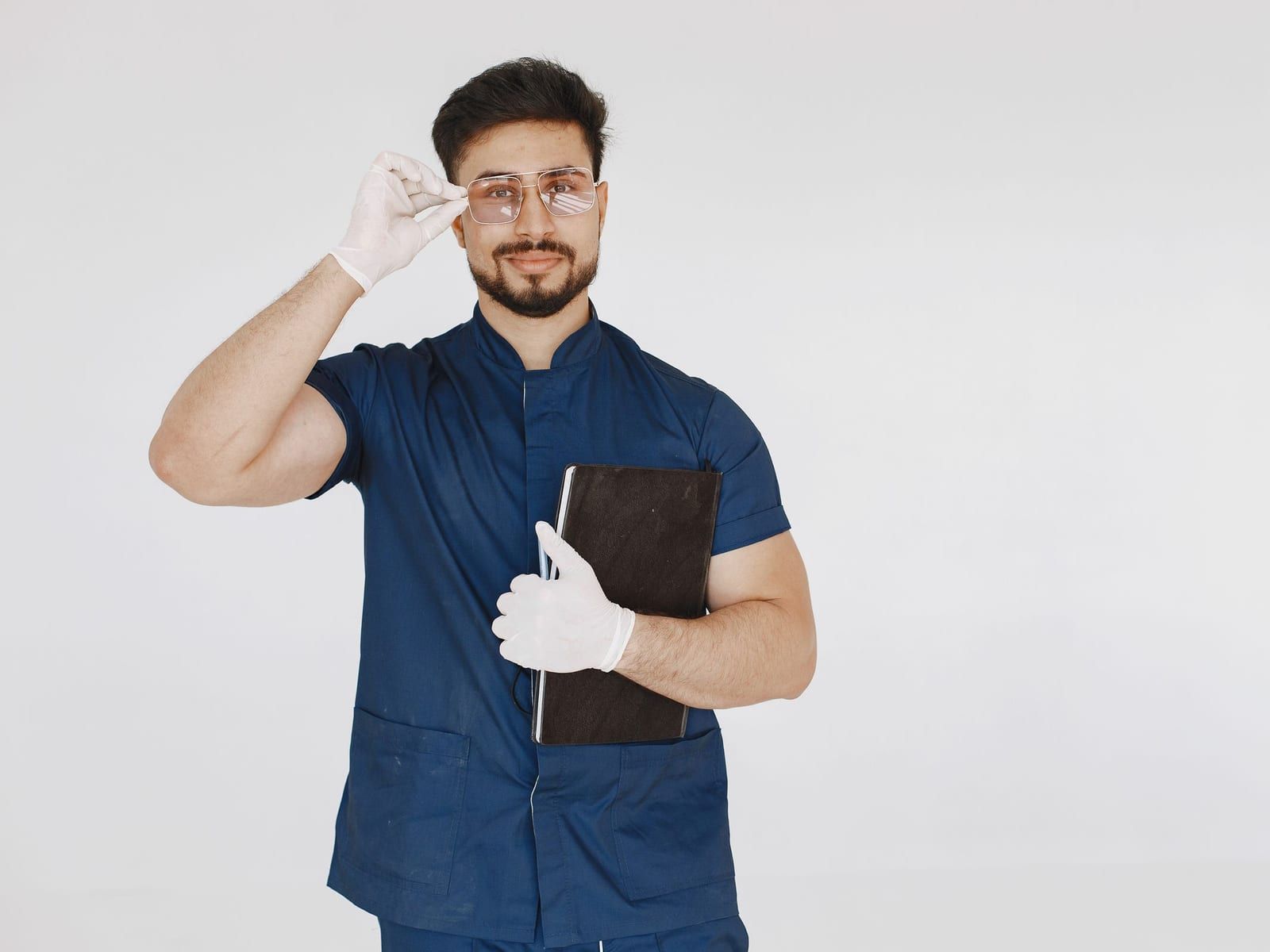 A bearded man in navy short-sleeved scrubs and white exam gloves adjusts his glasses with one hand and holds a black notebook against his chest with the other, against a plain white wall