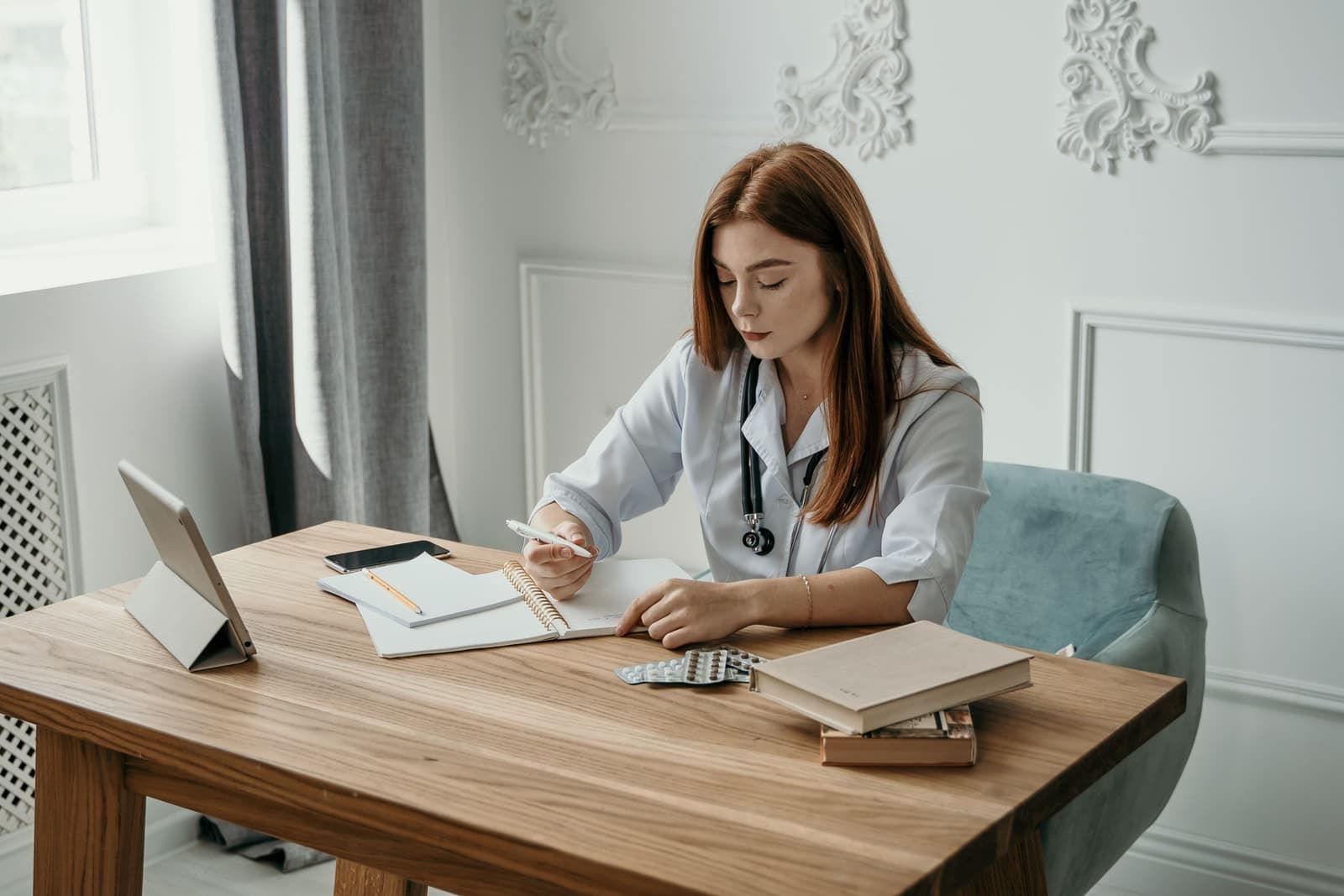 A clinician in a white coat and stethoscope taking notes at a wooden desk with a tablet, reference books, and blister packs of medication, the study side of PMHNP training