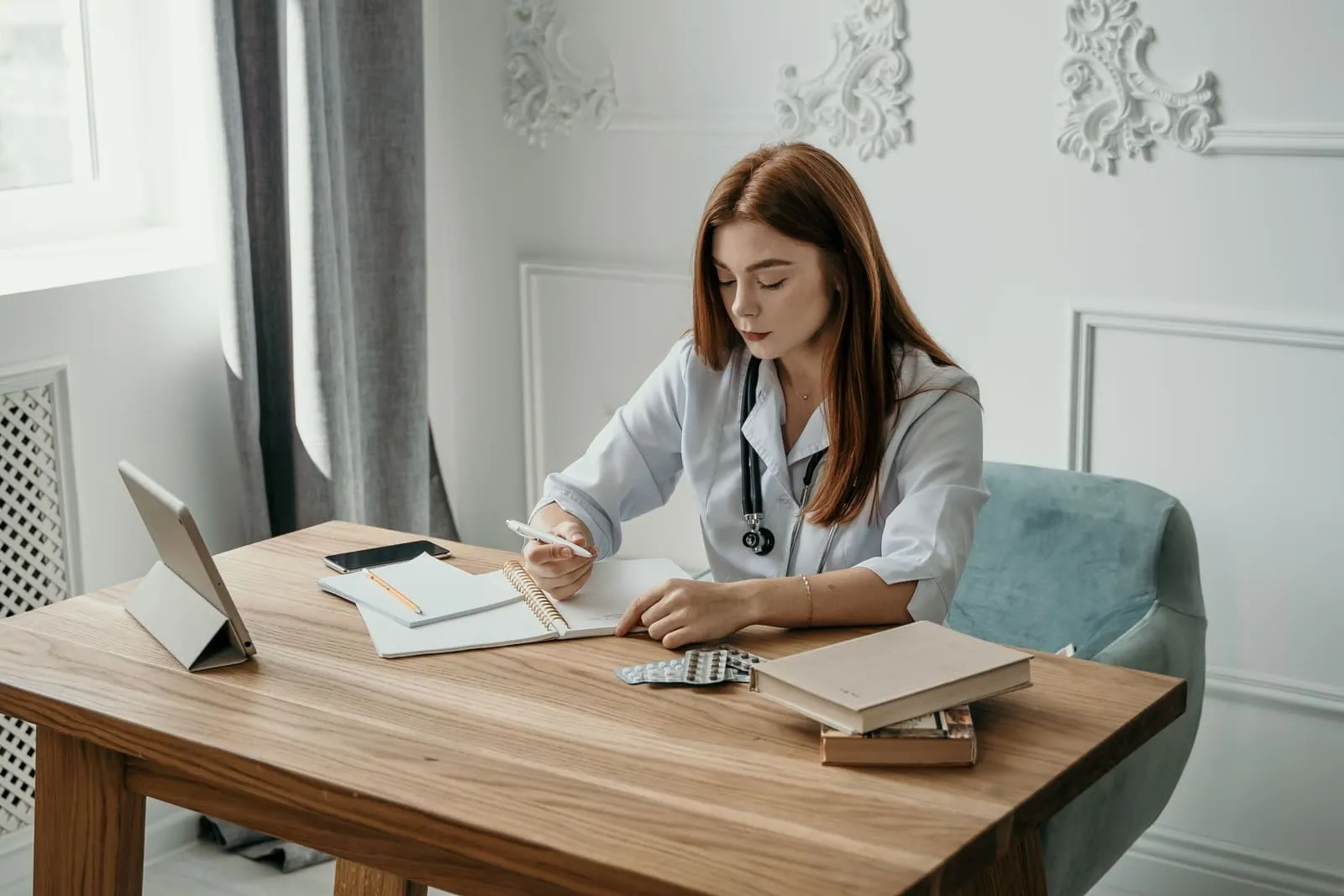 A clinician in a white coat and stethoscope taking notes at a wooden desk with a tablet, reference books, and blister packs of medication, the study side of PMHNP training