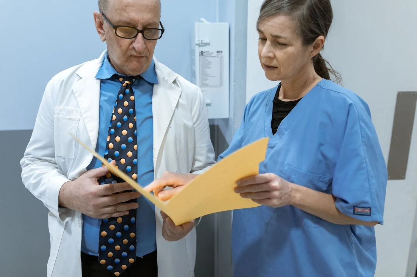 A nurse practitioner in scrubs sitting beside a student at a clinic desk, both looking at the same chart, the shape of a precepted clinical day