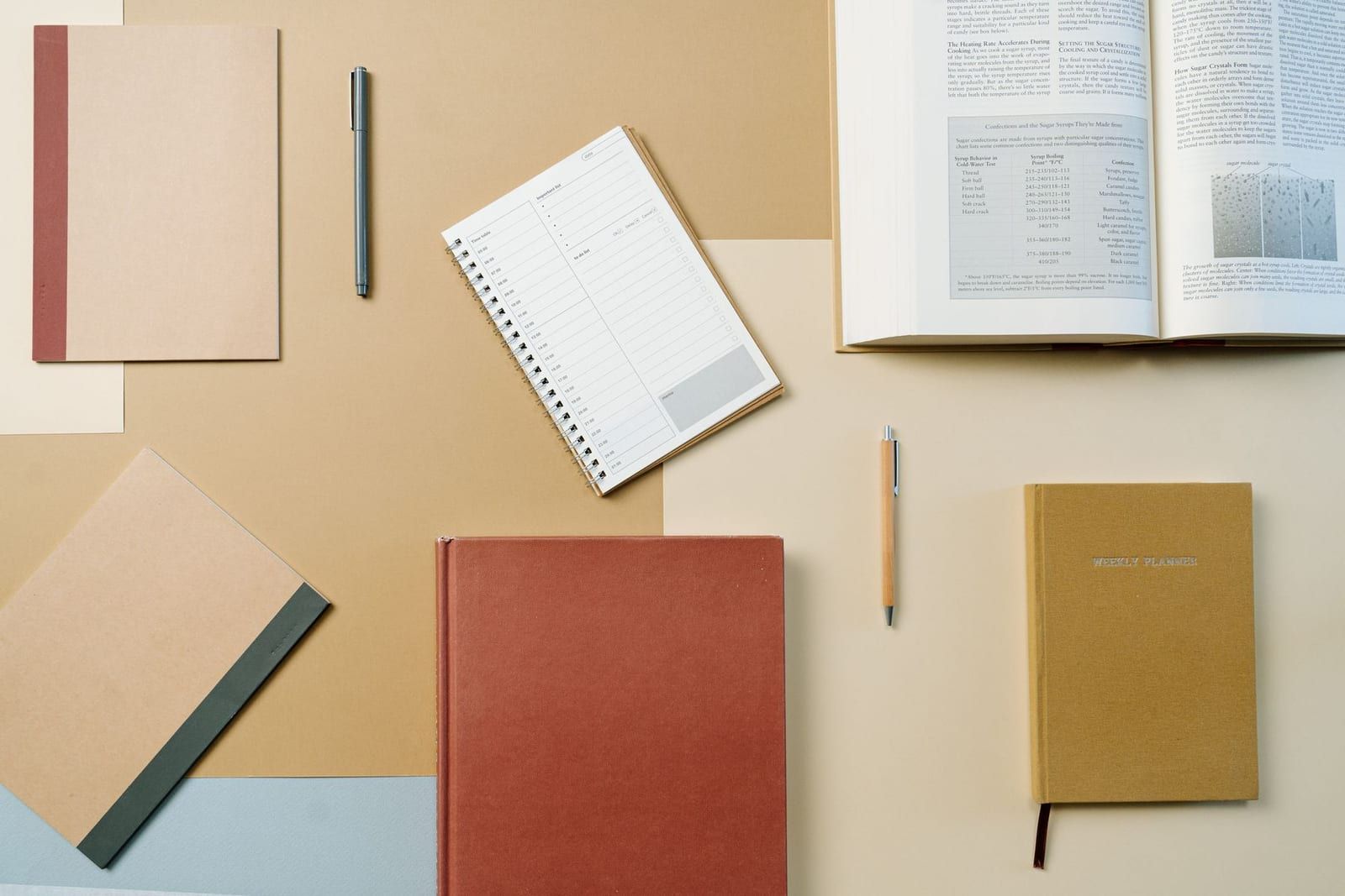Overhead view of a study desk in warm tan and terracotta tones, with stacked hardcover notebooks, an open reference book, a spiral daily planner, and two pens arranged on layered sheets of paper