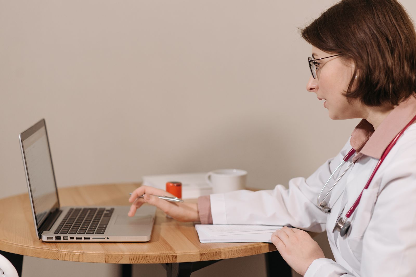 A clinician in a white coat with a red stethoscope working at a laptop on a round wooden table, a notebook, a prescriber stamp and a mug beside her