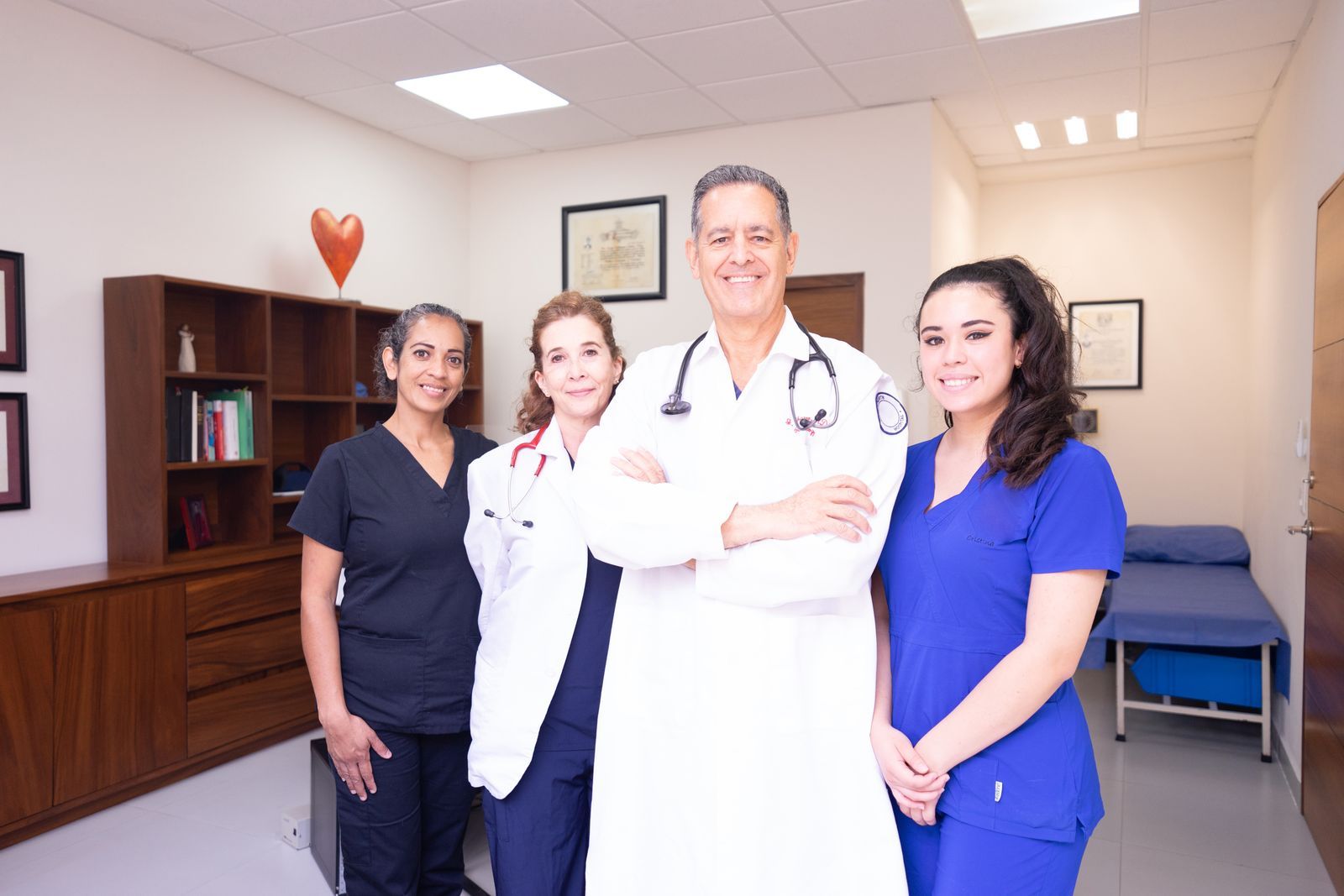 Four clinicians in a bright clinic consultation room, two in white coats with stethoscopes and two in scrubs, standing together in front of a bookshelf and framed certificates