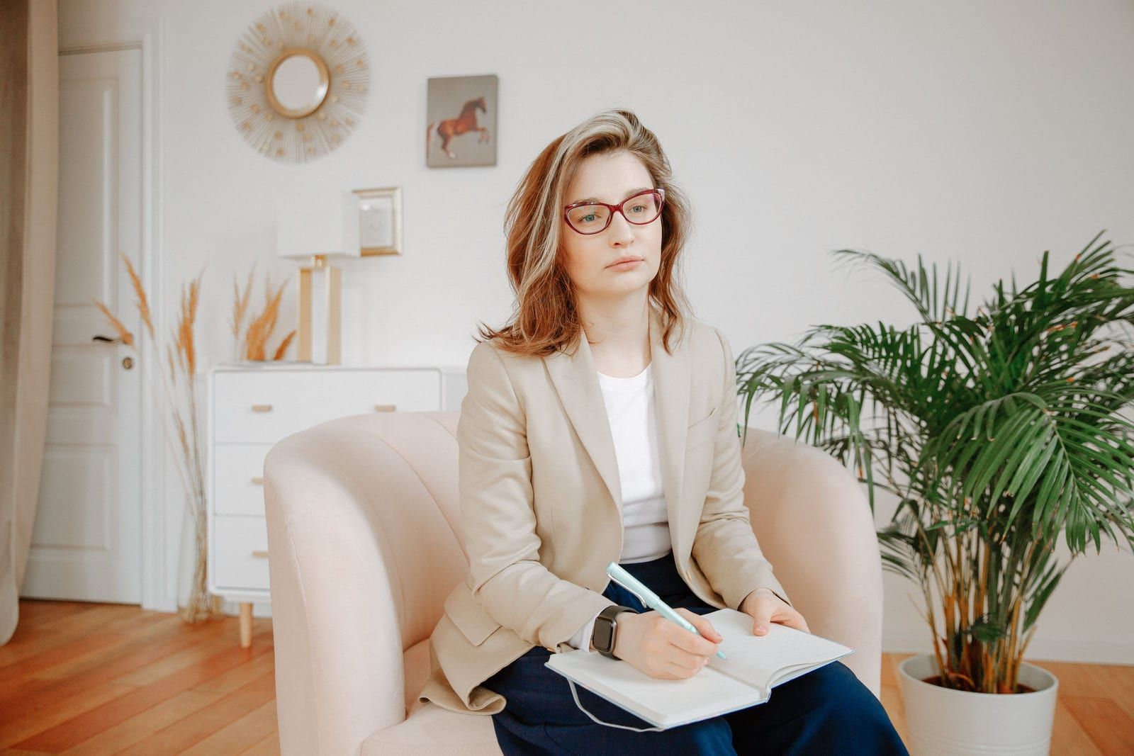 A clinician in a beige blazer and dark red glasses sitting in a pale armchair in a bright consulting room, an open notebook and pen resting on her lap