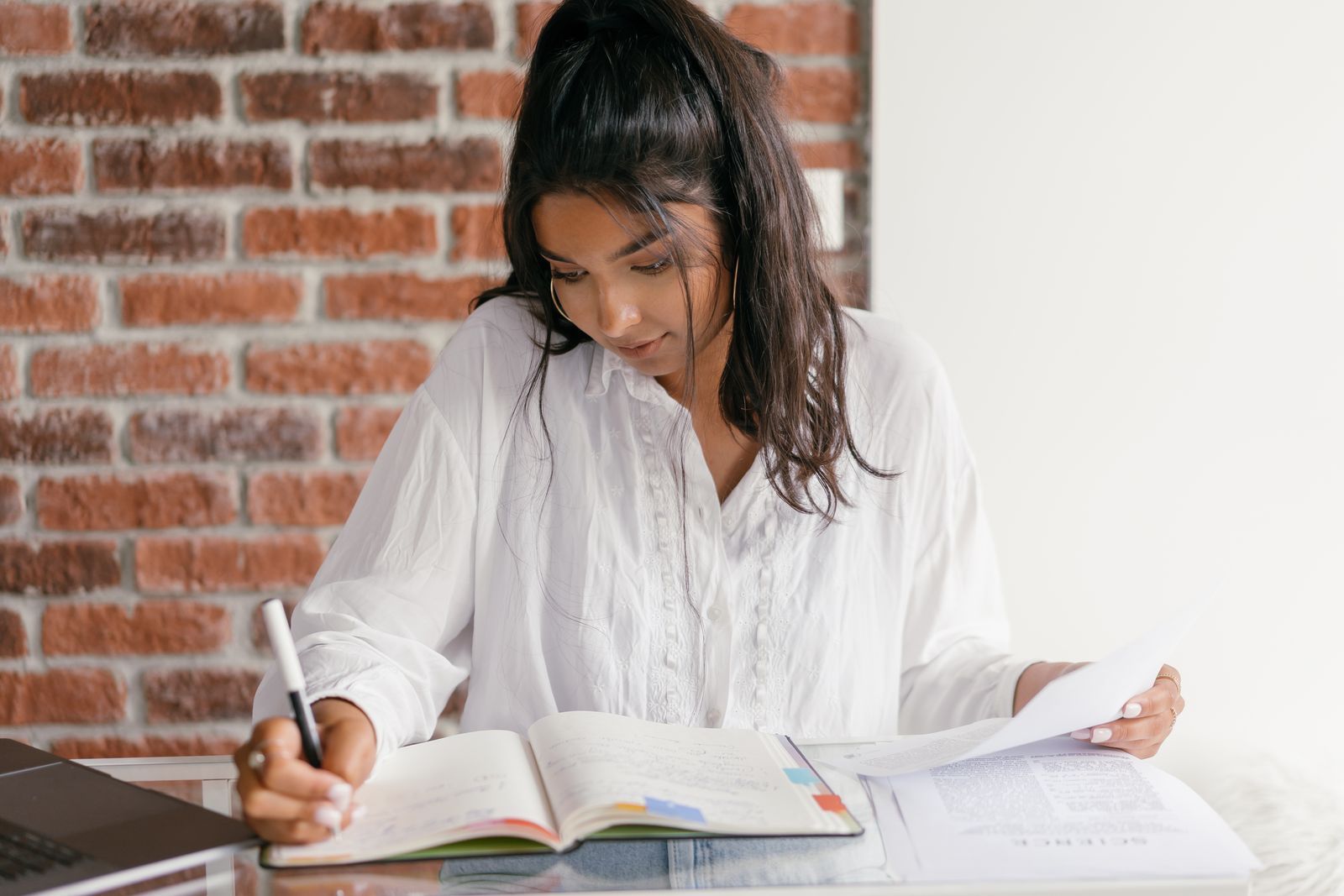 A student in a white blouse writing in a notebook at a desk against a brick wall, a printed form in her other hand and a laptop at the edge of the frame