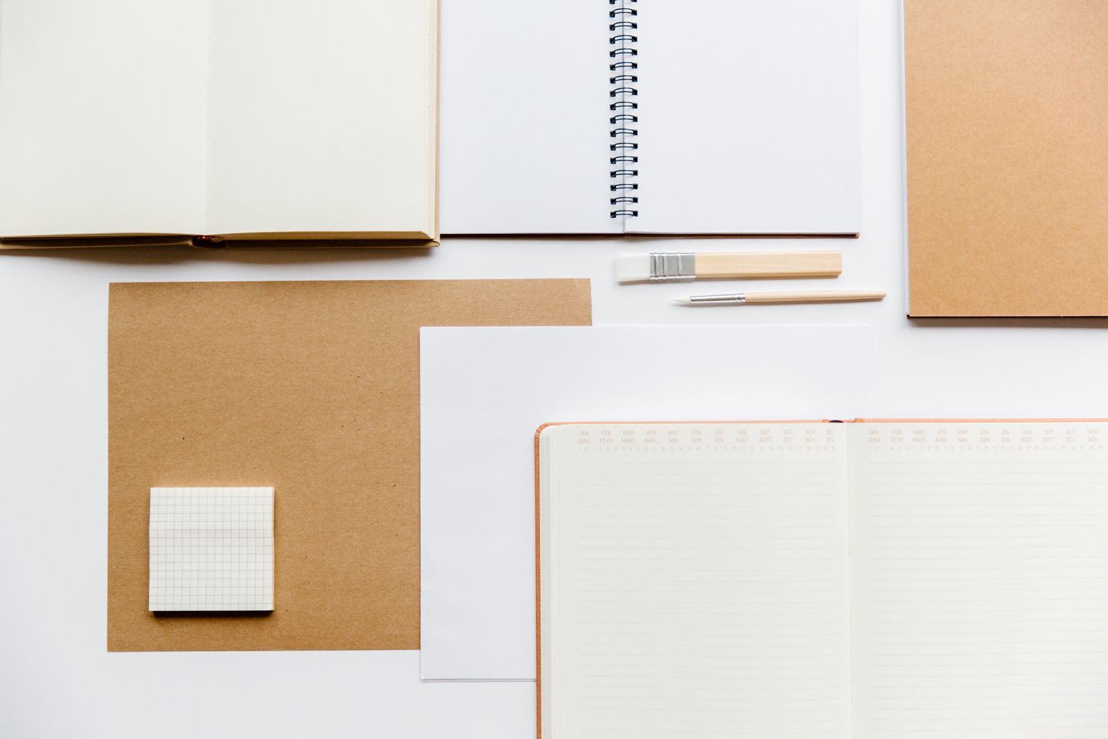 Overhead flat lay of open notebooks, a month-tabbed planner, kraft paper and pens arranged on a white desk.