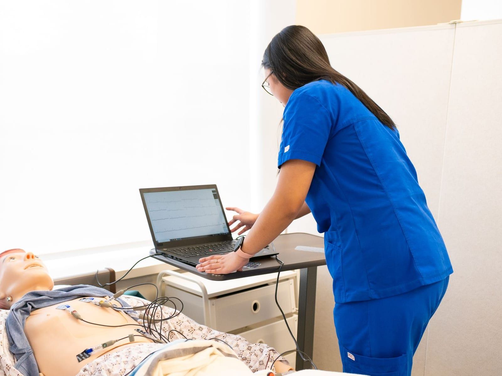 A nurse in royal blue scrubs leans over a laptop showing a heart rhythm tracing beside a patient simulation mannequin wired to monitor leads in a bright training room